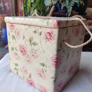 May include: A rectangular storage box with a quilted cream-colored exterior, adorned with pink rose floral patterns and green leaves. The box has a lid and rope handles for carrying. The overall design is vintage and decorative.