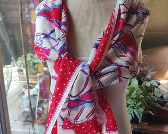 May include: A colorful scarf with a vibrant design, draped around a mannequin. The scarf features a mix of red, white, and blue patterns, with a polka dot border. The scarf is knotted at the front, showcasing its flowing fabric.