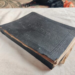 May include: A vintage, black, hardcover book with embossed lettering and decorative border details. The book's edges show signs of wear and age, with visible damage to the spine and corners. The book is resting on a white surface.