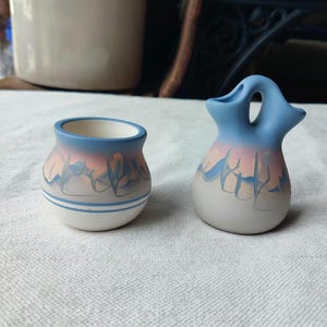 May include: Two small, hand-painted ceramic vases. One is a small pot with a blue rim and a design of blue and pink brushstrokes. The other is a vase with a handle, also with a blue rim and similar brushstroke design.
