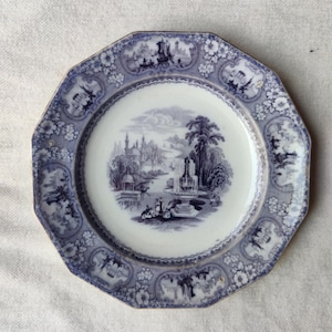 May include: An antique blue and white octagonal plate. The plate features a central landscape scene with a fountain and buildings, surrounded by a floral and architectural border. The plate has a gold rim.
