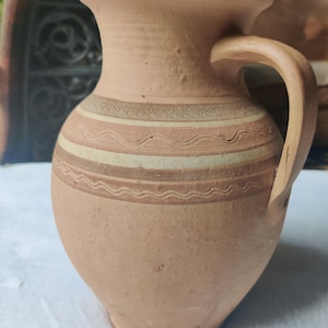 May include: A handmade, terracotta-colored ceramic pitcher with a handle. The pitcher features horizontal bands of tan, cream, and brown decorative patterns around the body. The pitcher has a rounded base and a flared rim.