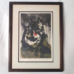 May include: A framed abstract print featuring dark, textured black ink strokes and muted olive green tones on a cream background. A single, small, vibrant red heart shape is positioned near the center of the composition, enclosed in a dark brown frame.