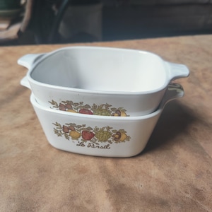 May include: Two white, square casserole dishes stacked on top of each other. Each dish has a decorative band of colorful fruits and vegetables. The words "Le Persil" are printed on the front of the bottom dish.