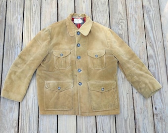 Peut inclure: Veste en velours côtelé beige avec quatre poches avant et des boutons bleus. La veste a un col et une doublure à carreaux visible au col. La veste est posée sur une surface en bois.