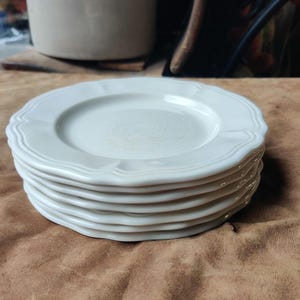 May include: A stack of white ceramic plates with scalloped edges. The plates have a simple, elegant design and are stacked on top of each other. The plates are a neutral color and would complement any table setting.