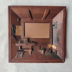 May include: A detailed wooden diorama depicting a workshop scene. The miniature room features a workbench, tools, a small stool, and a fireplace. The frame is made of dark wood, and the interior is intricately carved.