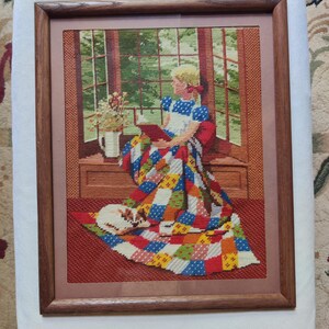 May include: A framed needlepoint tapestry depicting a person with blonde hair sitting in a window seat reading a book. The person is covered by a colorful patchwork quilt with a small cat resting on the floor nearby. A vase of flowers sits on the sill.
