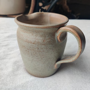 May include: A handmade ceramic mug with a rounded body and a curved handle. The mug has a light green and brown speckled glaze. The rim is slightly flared, and the interior is a lighter shade of brown. The mug is sitting on a white surface.