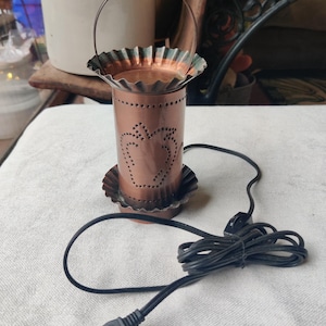 May include: A copper-colored electric candle warmer with a heart-shaped design. The warmer has a handle and a scalloped rim. A black power cord with a plug is connected to the warmer.