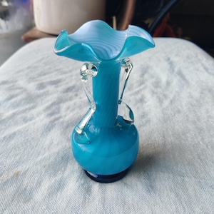 May include: A turquoise glass vase with a ruffled rim and clear glass handles. The vase has a round base and a bulbous body. The rim is a lighter shade of turquoise with white swirls. The vase is approximately 6 inches tall.