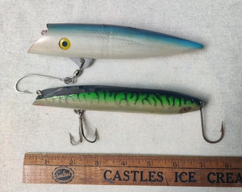 May include: Two fishing lures positioned above a wooden ruler measuring approximately 8 inches. The top lure is white and blue with a yellow eye. The bottom lure features a dark green and black mackerel-style pattern with two metal treble hooks.