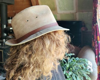 May include: A tan-colored fedora-style hat with a brown leather band. The hat has two small eyelets on the crown. The hat is worn by a person with curly, light brown hair. The hat appears to be made of a soft material.