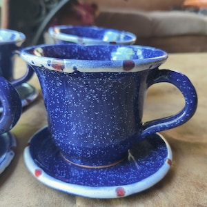 May include: A set of blue ceramic teacups and saucers. The cups and saucers are a deep blue with white speckles and a white rim with red and brown accents. The cups have a curved handle.