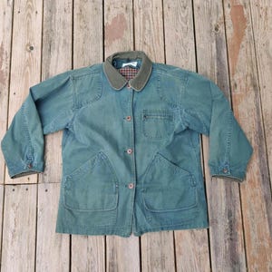 May include: A teal-colored jacket with a corduroy collar and button closure. The jacket has two large front pockets and a smaller chest pocket. The collar has a plaid pattern. The jacket is made of a durable material.