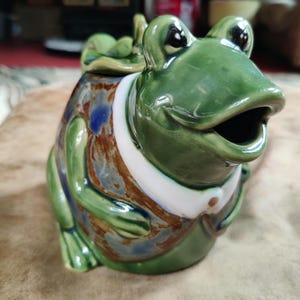 May include: A ceramic frog-shaped teapot with a green glaze. The frog has large eyes, an open mouth, and a white collar. The body is decorated with brown, blue, and white patterns. A small leaf-shaped lid sits on top.