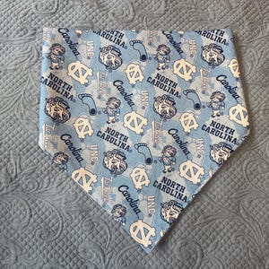 May include: A triangular pet bandana in light blue featuring a repeating pattern of the North Carolina Tar Heels mascot, the UNC logo, the text North Carolina, and blue footprints. The design includes various illustrations of the mascot.