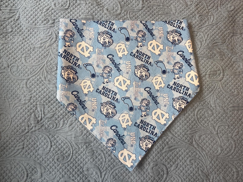 May include: A triangular pet bandana in light blue featuring a repeating pattern of the North Carolina Tar Heels mascot, the UNC logo, the text North Carolina, and blue footprints. The design includes various illustrations of the mascot.
