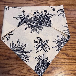 Puede incluir: Un pañuelo blanco con estampado de piña negro. El pañuelo tiene botones a presión negros en los bordes.