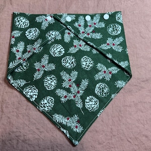 May include: A green bandana with a white pine cone and branch pattern. The bandana is folded into a triangle and secured with three white buttons. The design includes red berries.