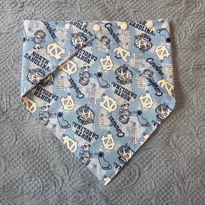 May include: A light blue triangular pet bandana featuring an all-over print of North Carolina Tar Heels logos, mascots, and footprints. The fabric is patterned with the text North Carolina, Carolina, and UNC, and includes four white plastic snap buttons.