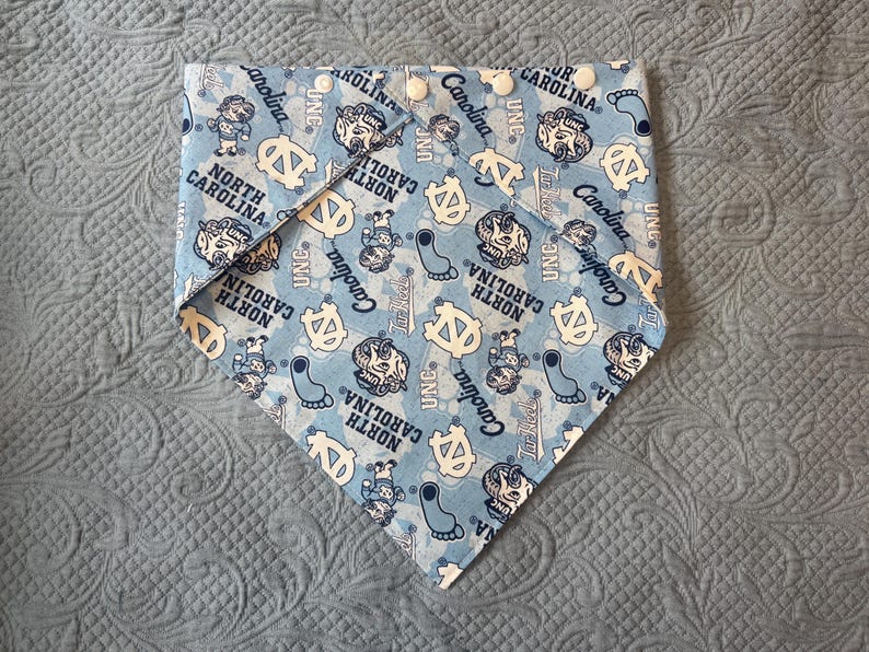 May include: A light blue triangular pet bandana featuring an all-over print of North Carolina Tar Heels logos, mascots, and footprints. The fabric is patterned with the text North Carolina, Carolina, and UNC, and includes four white plastic snap buttons.