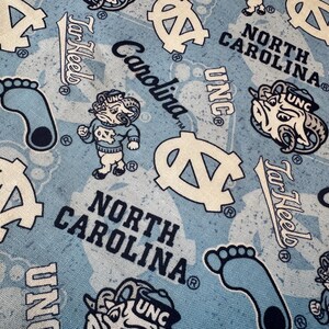 May include: Light blue fabric featuring a repeating pattern of North Carolina Tar Heels logos, including the ram mascot, the letters UNC, the word Carolina, the text North Carolina, and footprints in dark blue and white ink on a light blue background.