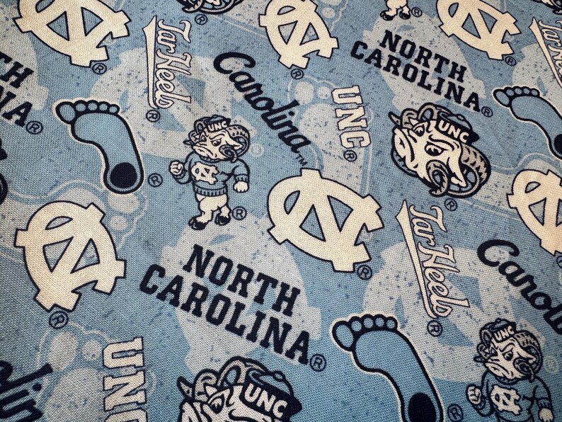 May include: Light blue fabric featuring a repeating pattern of North Carolina Tar Heels logos, including the ram mascot, the letters UNC, the word Carolina, the text North Carolina, and footprints in dark blue and white ink on a light blue background.