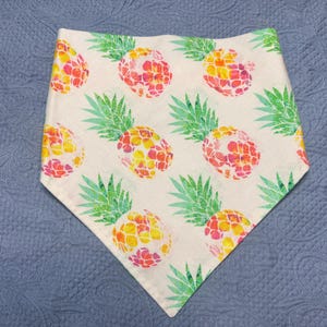 Puede incluir: Un pañuelo blanco con un patrón repetido de piñas coloridas. Las piñas tienen un efecto acuarela con tonos de amarillo, naranja y rosa. El pañuelo tiene forma triangular y está sobre un fondo azul.