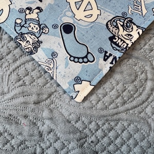 May include: A light blue fabric bandana featuring a repeating pattern of the University of North Carolina Tar Heels logo, a mascot character, and a footprint graphic in navy blue and white, finished with a stitched edge along the corner.