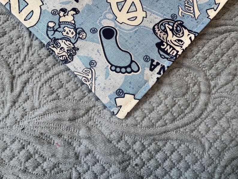 May include: A light blue fabric bandana featuring a repeating pattern of the University of North Carolina Tar Heels logo, a mascot character, and a footprint graphic in navy blue and white, finished with a stitched edge along the corner.