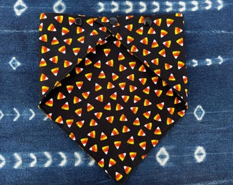 Puede incluir: Un pañuelo triangular negro para mascotas con un patrón repetitivo de caramelos de maíz en amarillo, naranja y blanco. El borde superior de la tela está doblado y asegurado con tres botones de presión de plástico negro para el cierre.