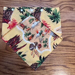 Puede incluir: Bandana amarilla con un estampado de isla tropical con palmeras, volcanes y pájaros. La bandana está doblada y asegurada con broches blancos. El diseño incluye acentos rojos, marrones y verdes.