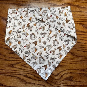 May include: A white bandana with a repeating pattern of brown and white chickens and chicken coops. The bandana has a triangular shape and is tied with two white buttons.
