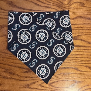 Può includere: Una bandana blu navy con un motivo ripetuto del logo dei Seattle Mariners e la lettera "S". Il logo è un design a rosa dei venti con accenti turchese e bianchi. La bandana è piegata a forma di triangolo.