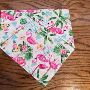 Puede incluir: Un pañuelo para perros con un estampado de flamencos tropicales. El pañuelo presenta flamencos rosas, flores tropicales y hojas verdes sobre un fondo blanco. Tiene cierres de botón a presión verdes.