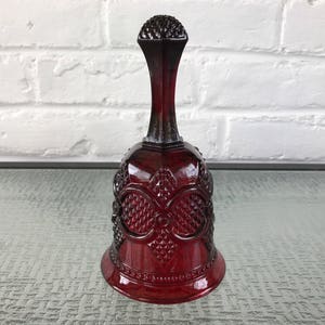 May include: A red glass bell with a decorative pattern. The bell has a wide base and a narrow top with a rounded handle.