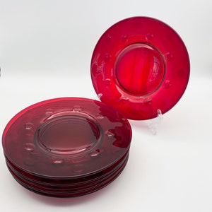 May include: A set of six red glass plates with a raised rim and a textured design. The plates are stacked on top of each other.