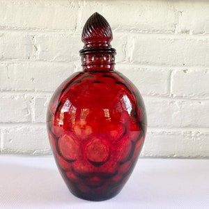 Puede incluir: Una jarra de vidrio rojo rubí con un cuerpo texturizado y un tapón decorativo. La jarra tiene forma redonda y es de vidrio transparente. El tapón tiene un diseño en espiral. Para licores o vino.