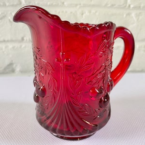 May include: A red glass pitcher with a cherry design. The pitcher has a handle and a textured surface.