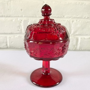 May include: A red glass candy dish with a lid. The dish is decorated with a grapevine pattern and has a pedestal base.