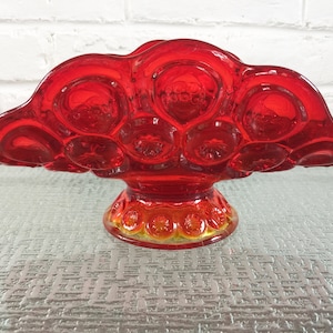 May include: A striking red glass dish with a starburst design. The dish features a scalloped rim and a pedestal base. The glass is a rich, transparent red, and the base has a yellow-orange tint.