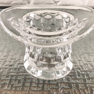 Puede incluir: Un portavelas de vidrio transparente con forma de sombrero de copa. El portavelas tiene un ala ancha y una base texturizada y facetada.