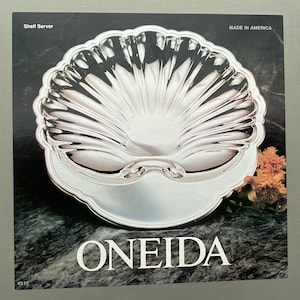 May include: A silver shell-shaped serving dish with a raised design. The dish is made by Oneida and says "Shell Server" and "Made in America" on the label.