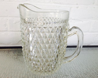 May include: A clear glass pitcher with a diamond pattern. The pitcher has a handle and a spout.