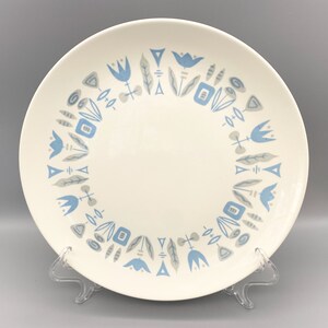 May include: A white ceramic dinner plate with a blue and gray floral pattern around the rim. The pattern features stylized flowers, leaves, and geometric shapes.