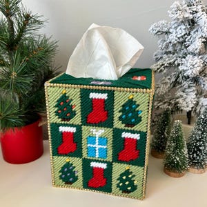 Puede incluir: Una funda para caja de pañuelos de color verde con temática navideña. La funda está decorada con calcetines rojos, árboles verdes y una caja de regalo azul. La funda tiene un ribete dorado.