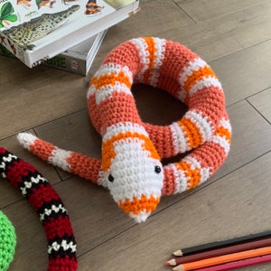 May include: A crocheted snake toy with orange and white stripes. The snake is curled up in a circle with its head facing the camera.