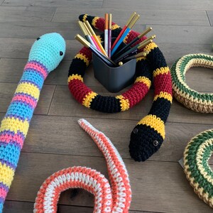 May include: A collection of crocheted snakes in various colors, including yellow, blue, pink, red, black, and green. The snakes are arranged on a wooden surface with a container of colorful pencils in the background.