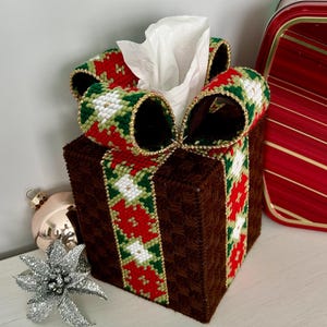 May include: A brown tissue box cover with a red, green, and white ribbon tied in a bow. The ribbon has a gold trim. The tissue box is sitting on a white surface.